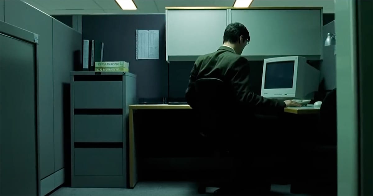 Thomas Anderson sitting alone in a dim office cubicle in a scene from "The Matrix."