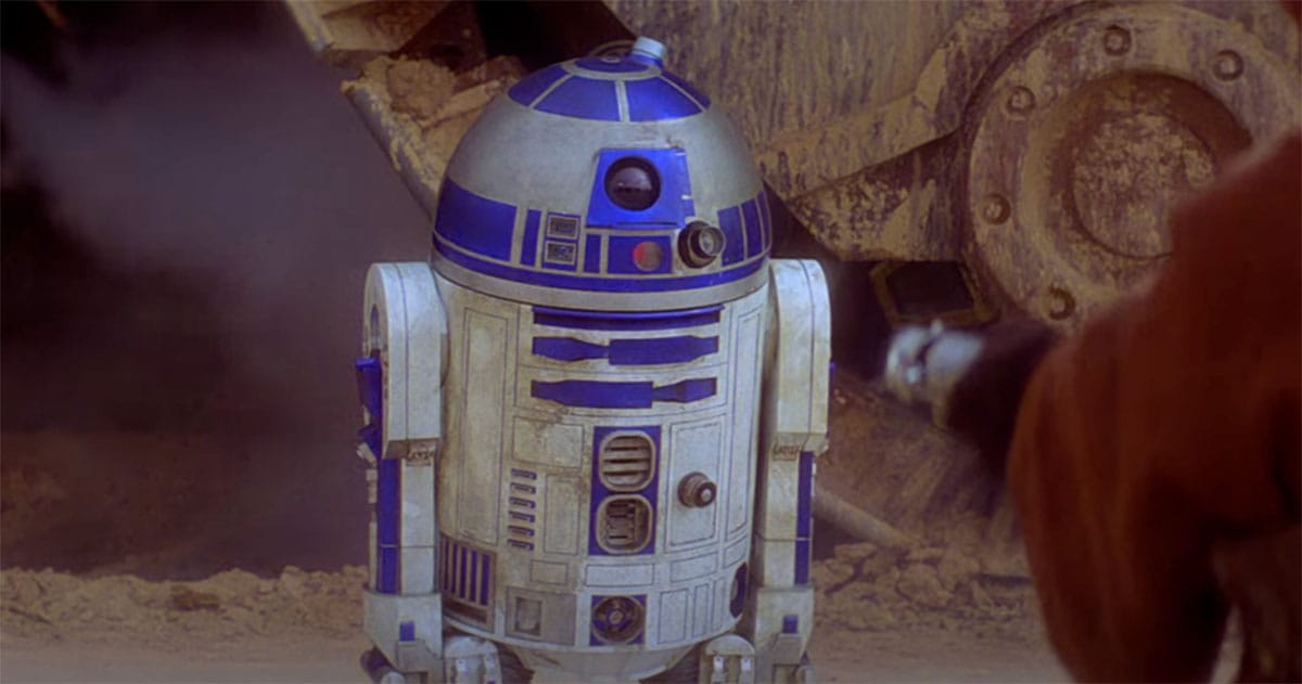 R2-D2 stands alone in a dusty setting from "Star Wars," with a dirt-encrusted machine and a robed figure partially visible in the background.