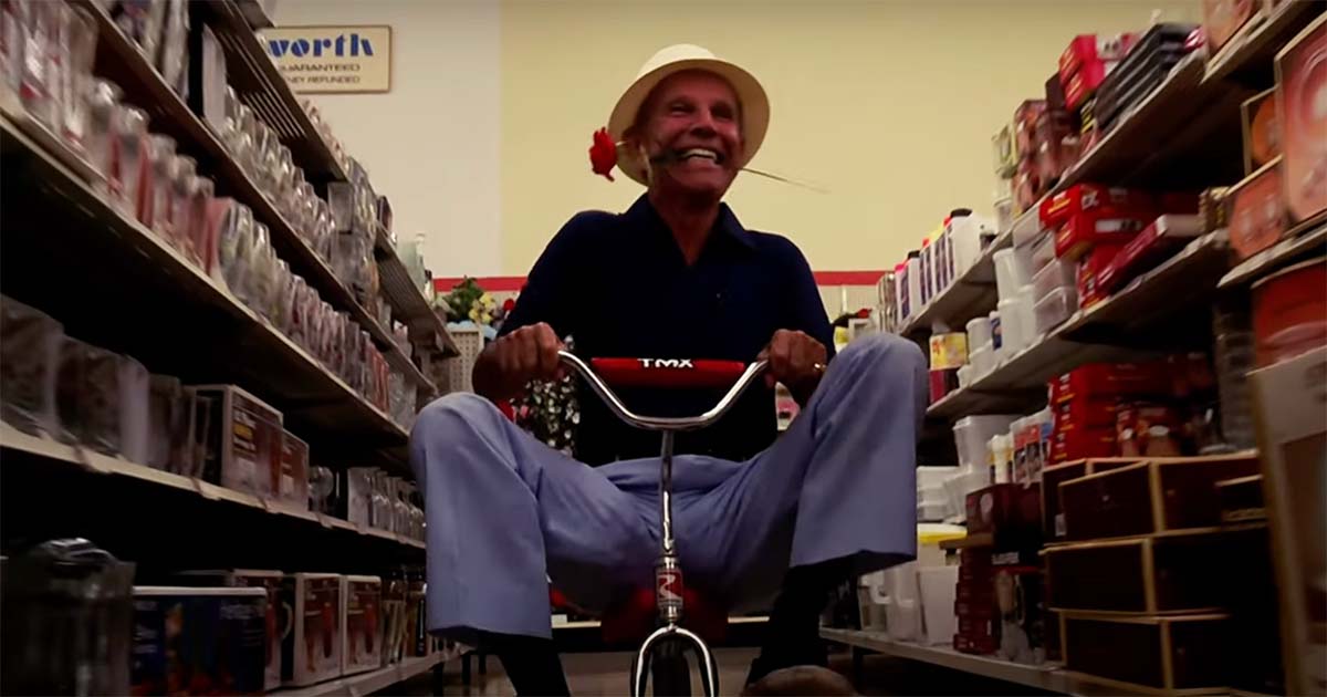 An elderly man rides a child's tricycle through a store aisle with a flower in his mouth.
