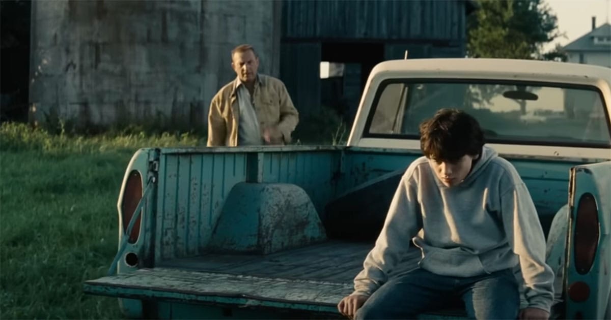 Young Clark Kent sits on the back of a pickup truck while Jonathan Kent approaches, capturing a quiet moment of moral guidance in "Man of Steel" on the Kent family farm.