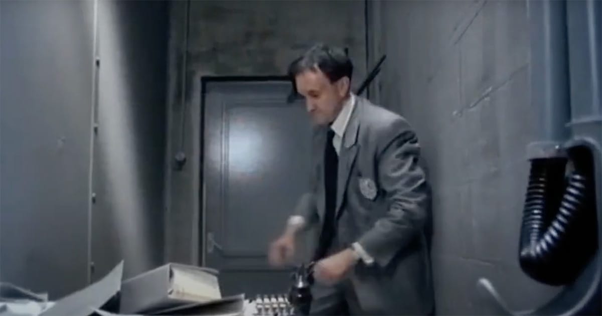 Sam Lowry stands in a narrow, cluttered office, holding an old-fashioned telephone beside a pile of paperwork, surrounded by gray walls and exposed ductwork in a scene from Terry Gilliam’s "Brazil" (1985).