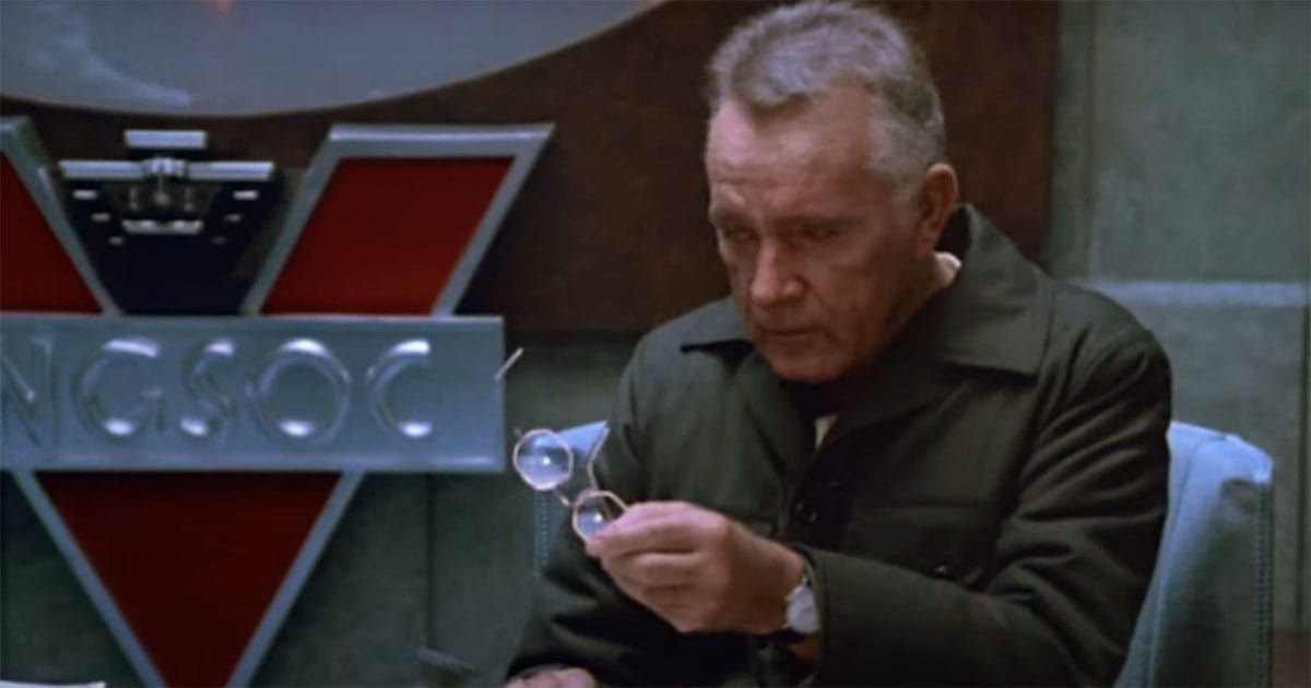 Richard Burton as O'Brien holding spectacles in an interrogation room under the INGSOC symbol in the 1984 film Nineteen Eighty-Four.