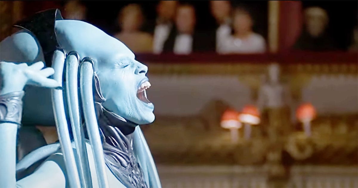Side profile of the Diva Plavalaguna singing with eyes closed and hand raised during her performance in "The Fifth Element," audience visible in the background.