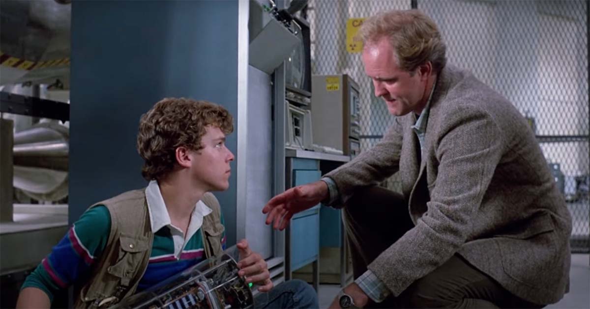 Paul sits on the lab floor holding a device while the scientist kneels and speaks to him seriously in “The Manhattan Project” (1986).