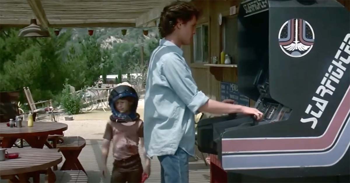 Alex Rogan playing the Starfighter arcade game outside while his younger brother Louis watches wearing a toy space helmet.