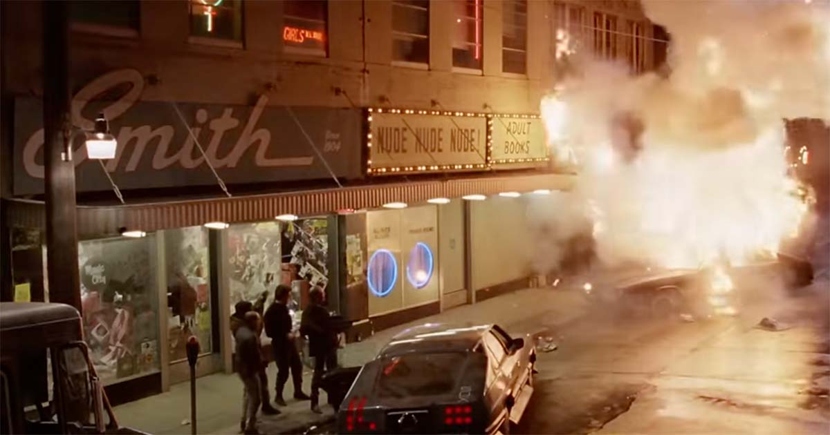 Scene from the 1987 film "RoboCop" showing Clarence Boddicker’s gang causing a massive car explosion outside a neon-lit Detroit storefront, representing the lawlessness and corruption that define the city’s decline.