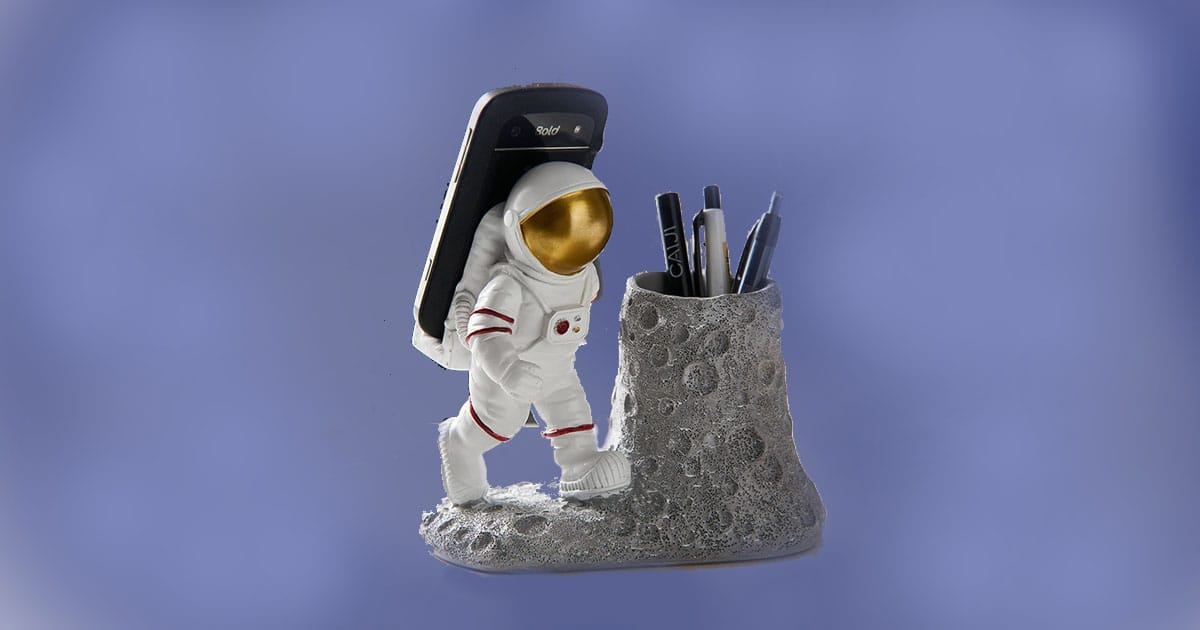 Astronaut-themed pen holder and phone stand featuring a white-suited astronaut with gold visor beside a crater-shaped container for pens.