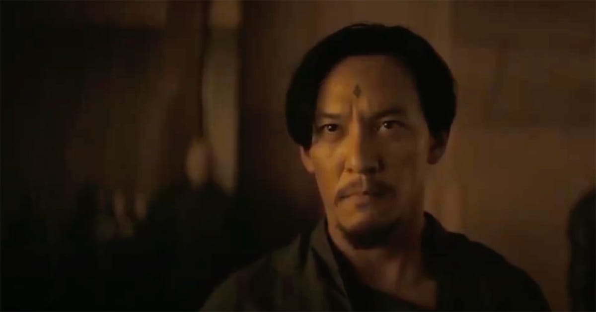 A man with a small diamond tattoo on his forehead looks ahead with grim focus under warm desert light.