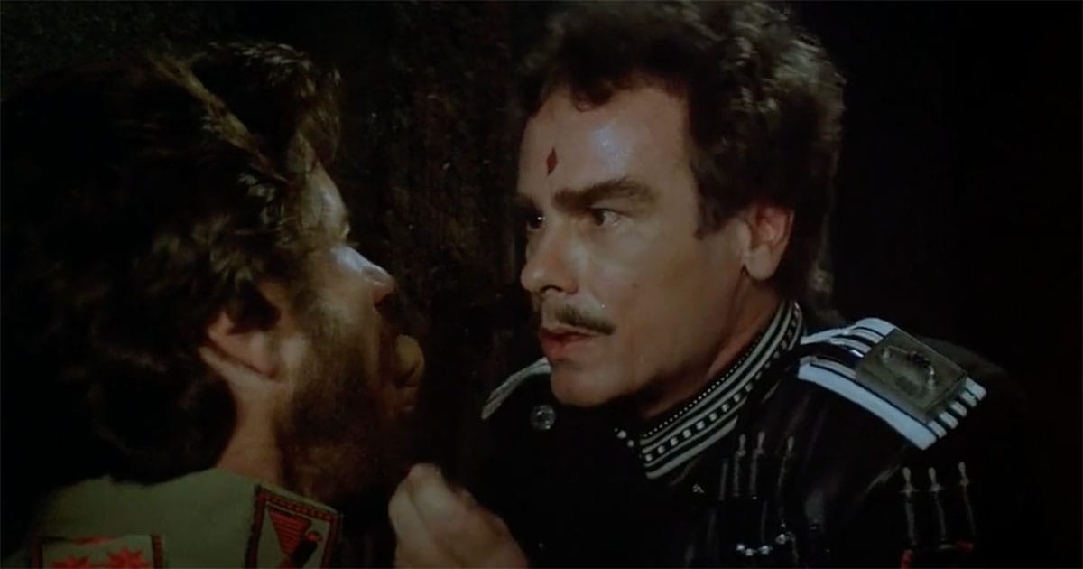 A man in a dark uniform with a small diamond mark on his forehead stares intensely at another man in a dim chamber.