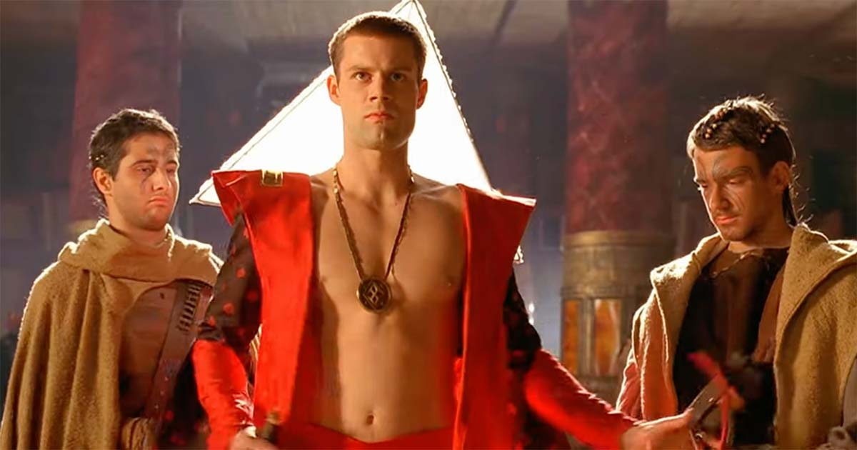 Actor Matt Keeslar as Feyd-Rautha stands in a red ceremonial robe between attendants in a large hall.