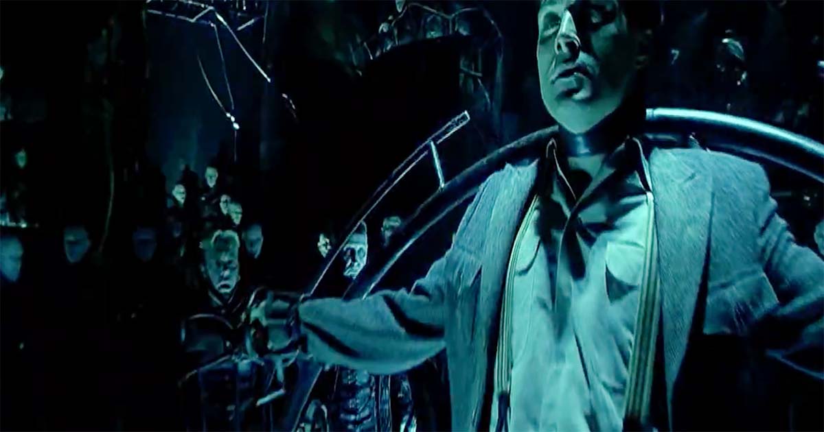 A man hangs restrained before a crowd of pale figures in a dark chamber from “Dark City.”