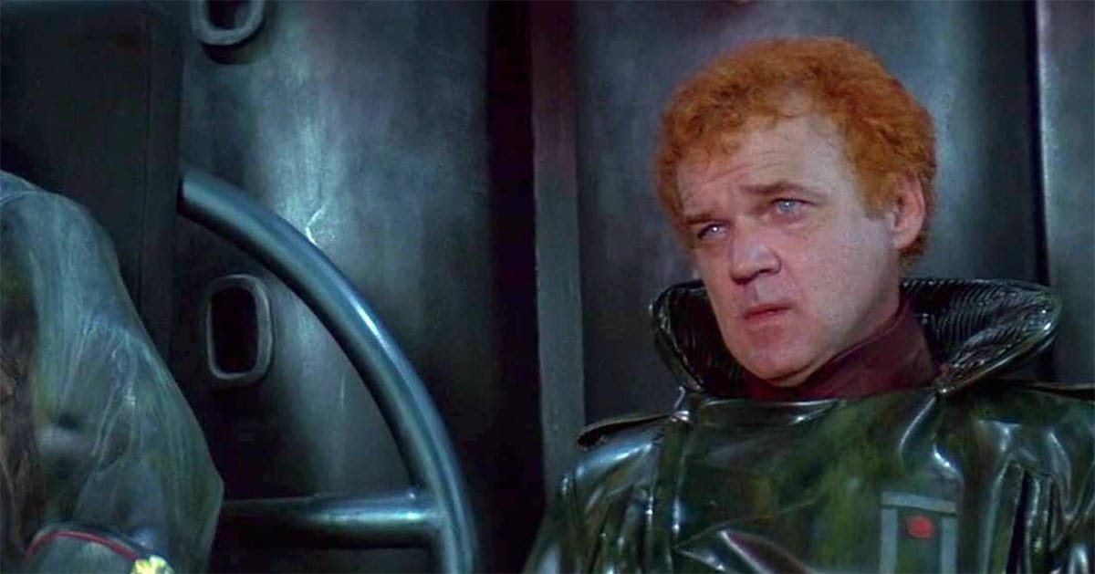 A man in glossy black armor sits before a dark wall, expression wary and tense.