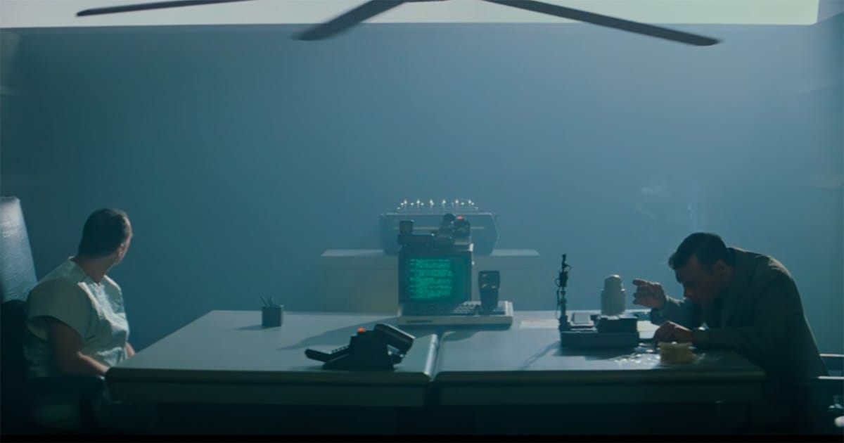 Two men sit across a table in a smoky room with testing equipment between them.