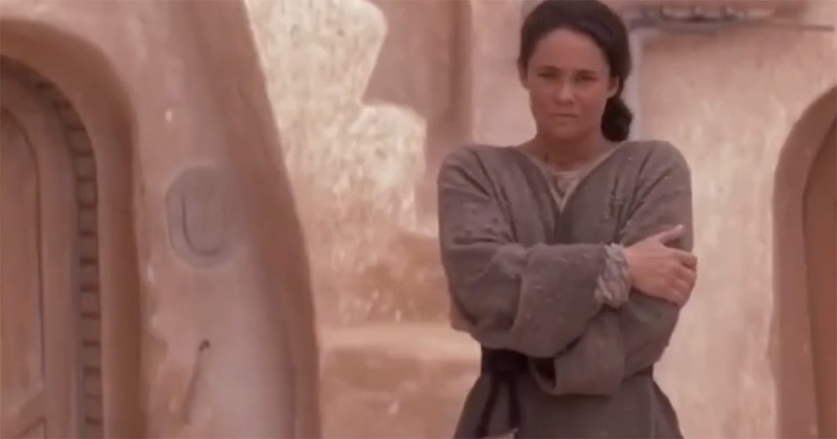 Shmi Skywalker standing outside her home with her arms folded in a moment of quiet reflection.