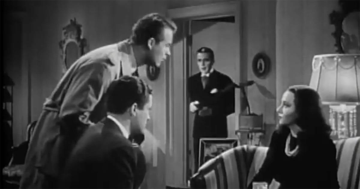 Three people speak in a living room while a man stands in the doorway watching in a scene from “The Return of Doctor X.”