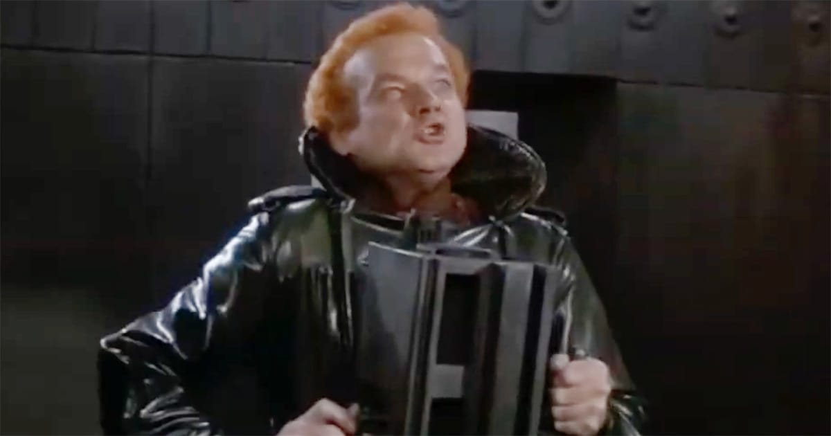 A Harkonnen guard captain grips a small box-like instrument close to his chest, his face tense under harsh industrial lighting.