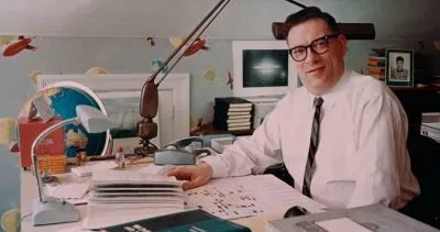 Isaac Asimov seated at his writing desk, wearing glasses and a tie, surrounded by papers and books in his study.