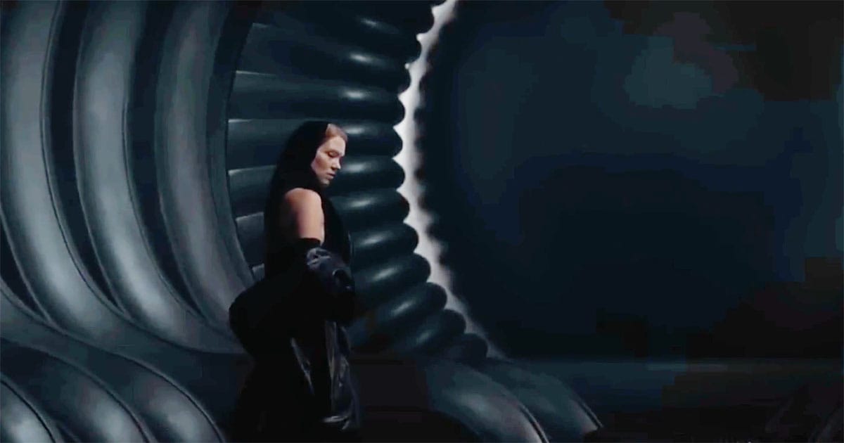 Lady Margot Fenring stands alone in a vast, curved interior corridor, her dark Bene Gesserit attire contrasting with the imposing imperial architecture.