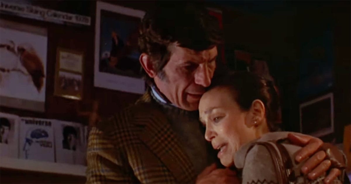 Man and woman embracing calmly indoors, appearing serene and emotionally distant in a scene from a 1970s science fiction film.