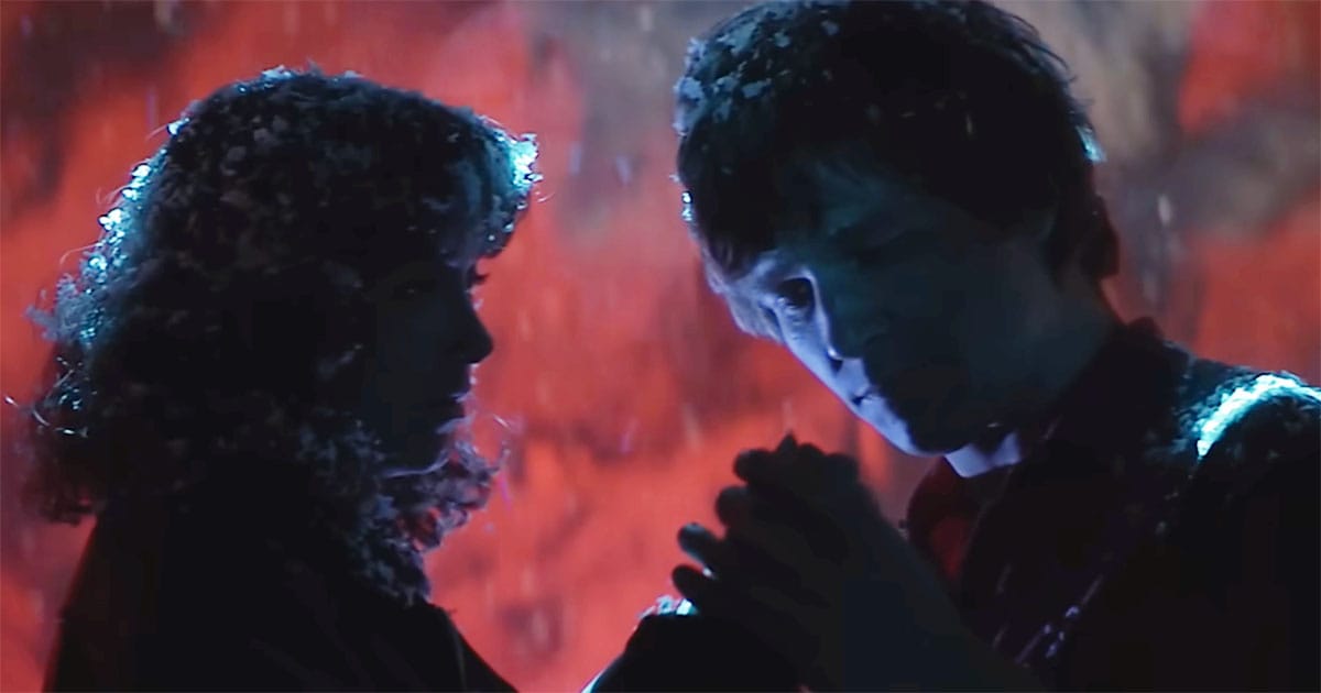 Two figures stand facing one another in falling snow and red light, hands gently clasped, sharing a quiet moment of trust and change.