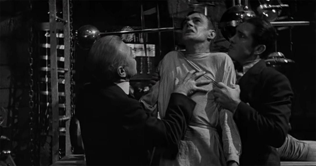 Laboratory scene from the 1931 film Frankenstein showing a man being held upright by two others amid scientific equipment.