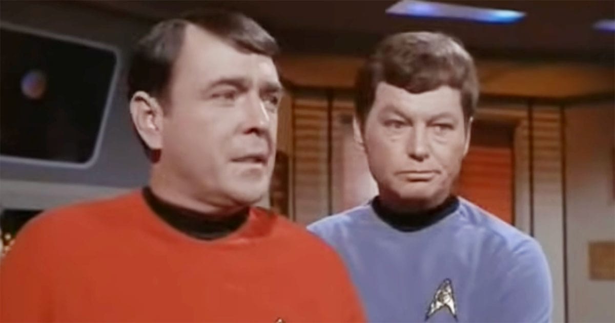 Young James Doohan as Scotty in red engineering uniform standing beside Dr McCoy on the Enterprise bridge in "Star Trek"