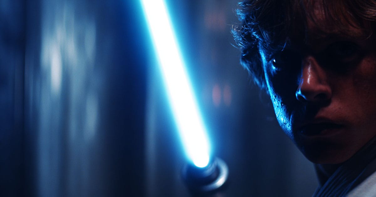 Close-up of a blue lightsaber igniting in a dark corridor, casting bright light across a focused young Jedi’s face in "Star Wars" (1977).