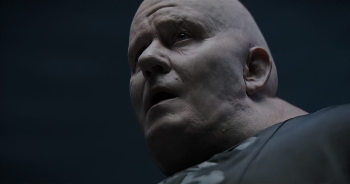 Close-up of a pale, heavyset man with a smooth bald head, gazing upward in dim light, his expression calm and commanding.