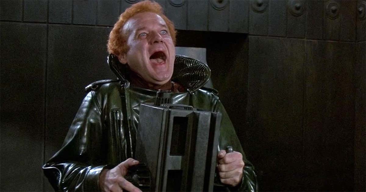 A uniformed guard captain with red hair holds a device and laughs inside a dark metal chamber.