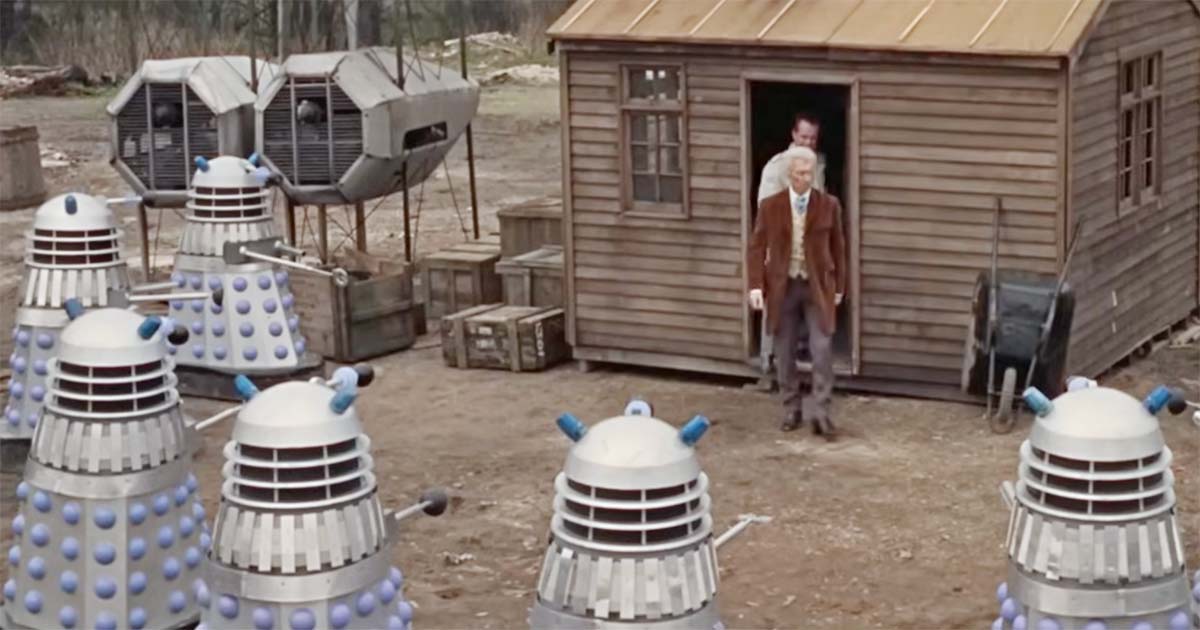 Dr. Who steps out of a wooden shed surrounded by Daleks in the 1965 film Doctor Who and the Daleks, showing mechanical aliens in a rural outdoor setting.