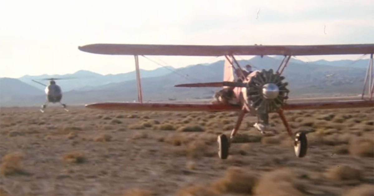 A vintage biplane flying low over a desert while a military helicopter pursues it in a climactic chase scene from the 1977 film Capricorn One.