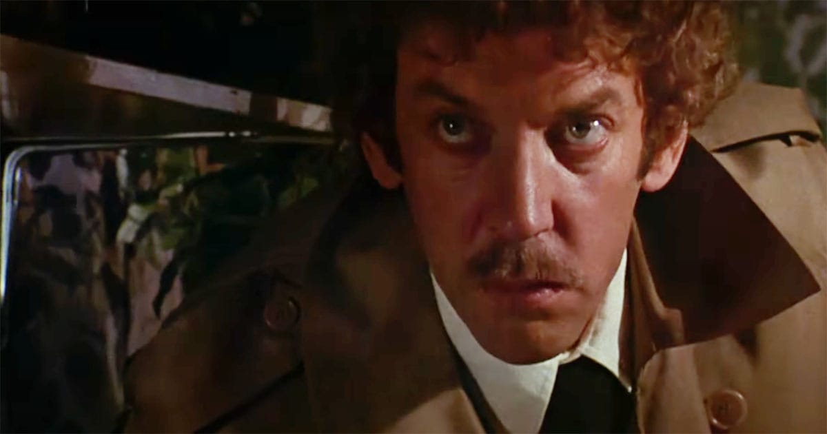 Close-up from a 1970s science fiction film showing a tense man in a trench coat sitting in a car, eyes alert and expression wary.