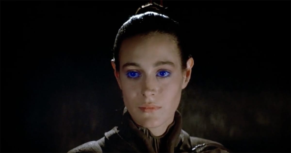 Close-up of Chani from "Dune" 1984 with blue eyes and a calm expression, wearing a stillsuit against a dark background.