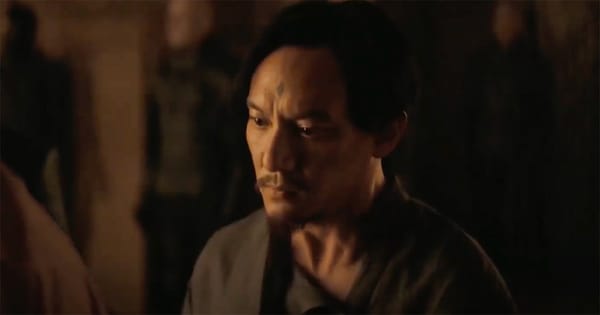 A weary man with a diamond mark on his forehead stands in dim amber light, his expression caught between sorrow and resolve.