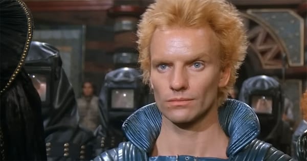 Actor Sting as Feyd-Rautha in blue armor, facing forward under harsh lighting with Harkonnen guards behind him.