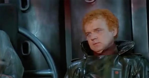 A red-haired Harkonnen officer in dark armor sits against a metallic wall, watching cautiously in a dim industrial chamber.