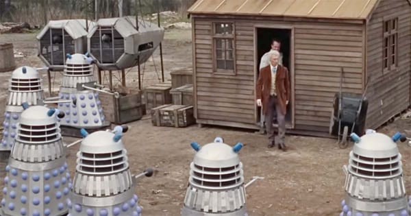 Dr. Who steps out of a wooden shed surrounded by Daleks in the 1965 film Doctor Who and the Daleks, showing mechanical aliens in a rural outdoor setting.