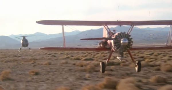 A vintage biplane flying low over a desert while a military helicopter pursues it in a climactic chase scene from the 1977 film Capricorn One.