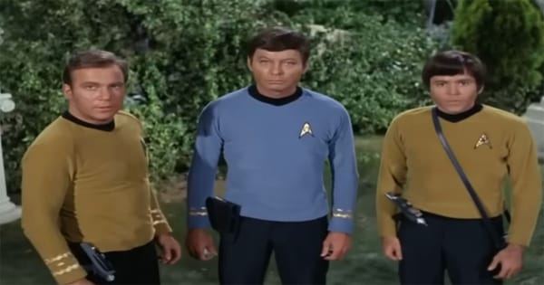 Three Star Trek original series officers standing outdoors with visible phasers and handheld equipment, wearing gold and blue uniforms during a field mission.
